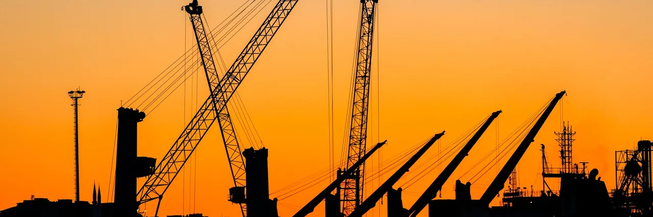 Silhouettes of tall industrial cranes against a vivid orange sunset sky
